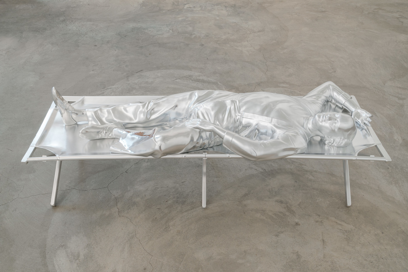 Life size horizontal statue of a man laying on a stretcher-like structure in a gallery space