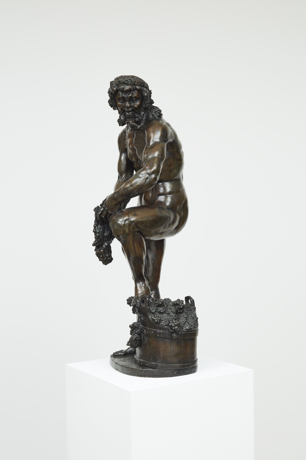Statuette of a realistically depicted bacchic man standing in a relaxed position with his foot perched on a basket filled with grapes