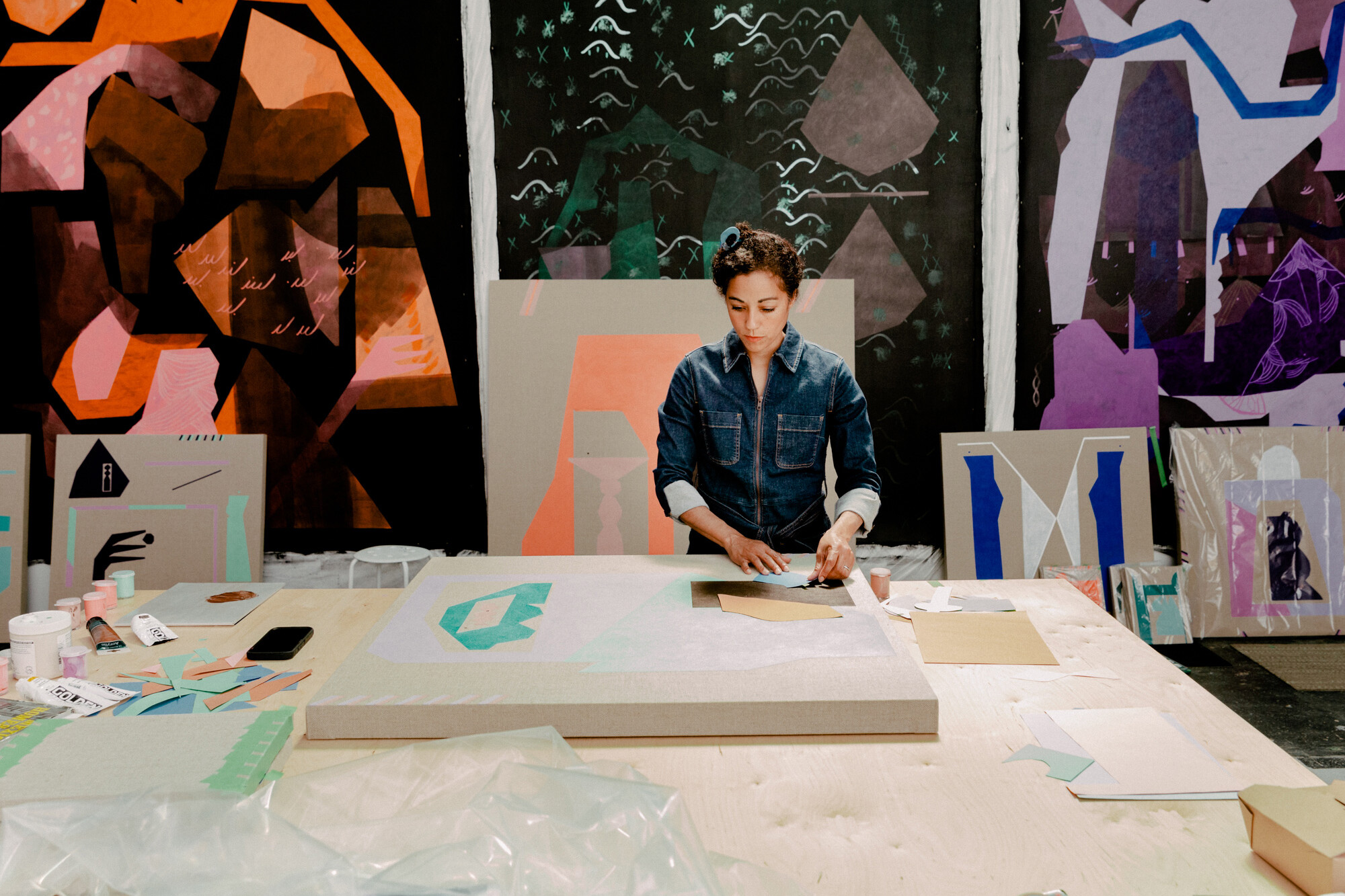 A woman in a denim jumpsuit in a studio. She is working on a collage on the table in front of her,...
