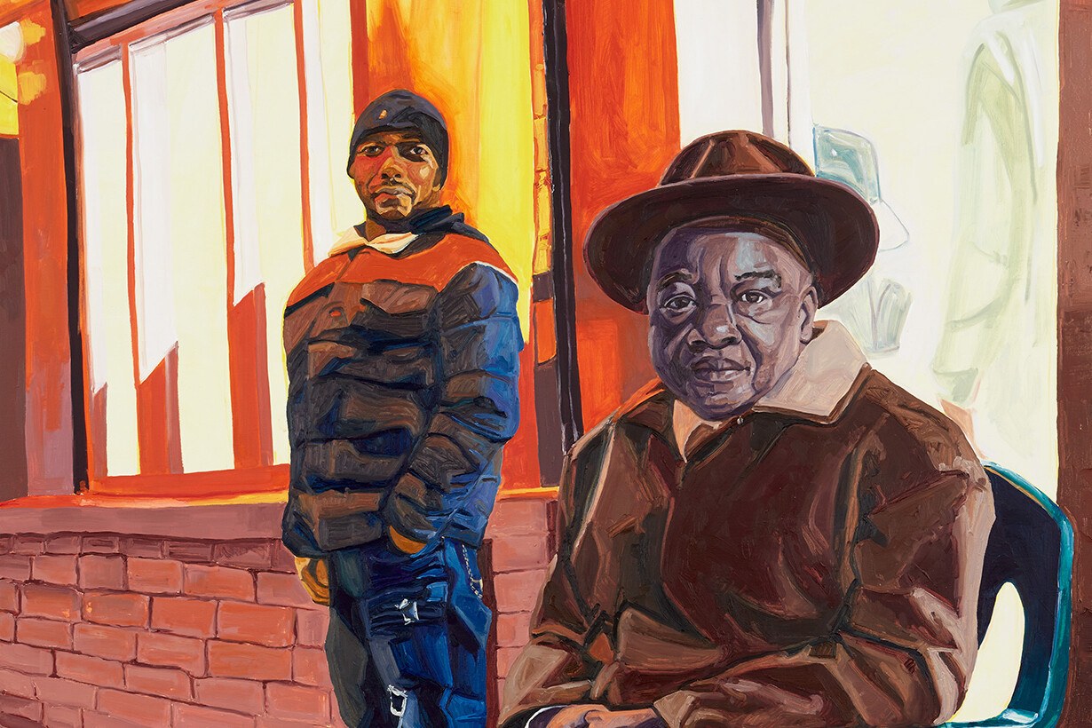 Man in hat seated in chair with younger man standing behind him.